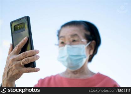 Happy Asian elderly wearing a face mask and answering a Video Call Using her Smartphone in the park and chatting with family, Technology and older people concept.