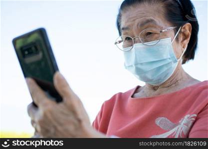 Happy Asian elderly wearing a face mask and answering a Video Call Using her Smartphone in the park and chatting with family, Technology and older people concept.