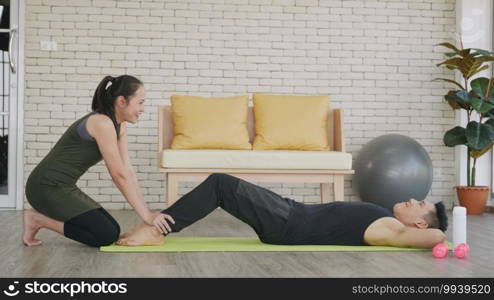 Happy Asian family couple husband and wife healthy trainer workout home in the living room. Fit man lifestyle doing sit up fitness exercise at home on floor with girlfriend support helping boyfriend