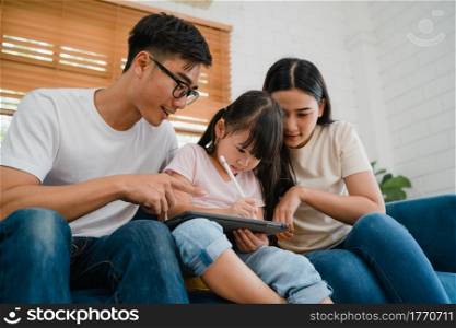 Happy Asian family dad, mom and daughter using computer tablet technology sitting sofa in living room at house. Self-isolation, stay at home, social distancing, quarantine for coronavirus prevention.