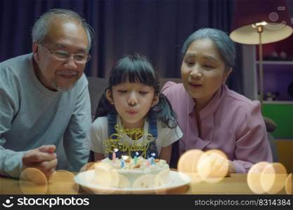 Happy Asian family grandparents and mother celebrating enjoy birthday party with cake of granddaughter Blowing candle at home in night time, Birthday surprise concept
