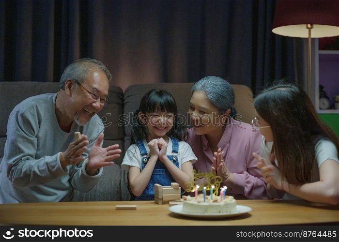 Happy Asian family grandparents and mother celebrating enjoy singing birthday song party with cake of granddaughter blowing candle at home in night time, Birthday surprise concept