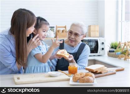 Happy Asian family preparing breakfast together. Grandmother, mother and grandchild girl spreading fruit jam on sliced bread and having fun in kitchen at home. Homemade food and little helper
