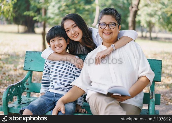 Happy Asian family with mother Grandmother and son sitting on the bench while the mother hugged them both in the park. The concept of lifestyle in a family holiday
