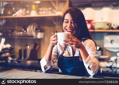 Happy Asian female barista holding cup of coffee and tasting brewed coffee from herself with cafe restaurant background. Waitress at cafeteria. Food and drink. People lifestyle and occupation concept.