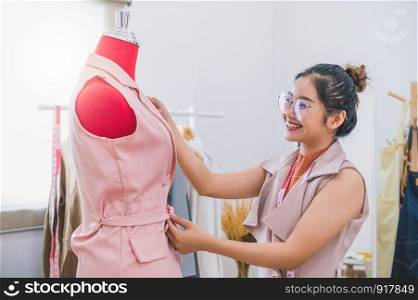 Happy Asian female fashion designer girl making fit on the formal dress uniform clothes on mannequin model. Fashion designer stylish showroom. Sewing and tailor concept. Creative dressmaker stylist.