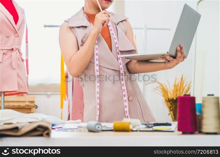 Happy Asian female fashion designer working in clothing shop studio and using laptop computer to contact customer. Woman creating design new collection . Tailor sewing. People lifestyle concept