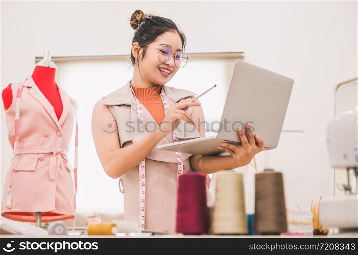 Happy Asian female fashion designer working in clothing shop studio and using laptop computer to contact customer. Woman creating design new collection . Tailor sewing. People lifestyle concept