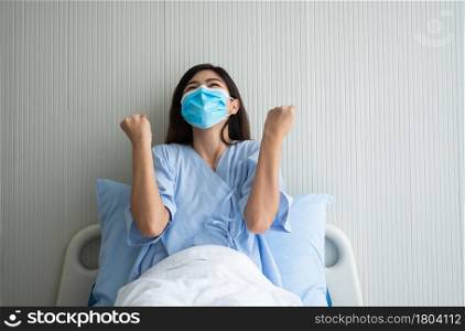 Happy Asian female patient wearing a mask, lies on the bed, and I raised an arms and was delighted for show confidence in treatment. Concept of believe in treatment And insurance coverage