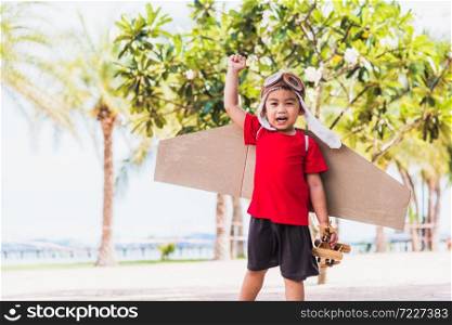 Happy Asian funny child or kid little boy smile wear pilot hat and goggles play toy cardboard airplane wing flying against summer sky cloud on trees garden background, Startup freedom concept