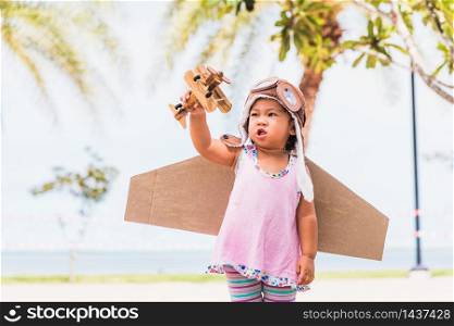 Happy Asian funny child or kid little girl smile wear pilot hat and goggles play toy cardboard airplane wing flying against summer sky cloud on trees garden background, Startup freedom concept