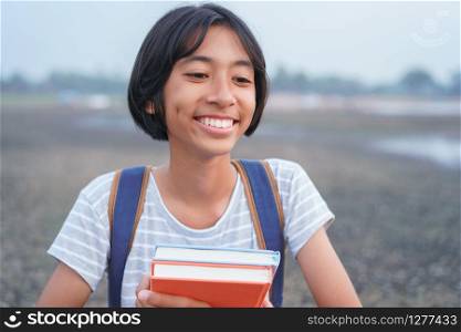 Happy Asian girl smile on face and laugh while standing Amid nature in the morning, Asia child hold book and backpack on blurred background. The schoolgirl came to study field trip and learned outdoor