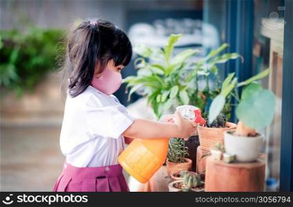 Happy Asian Girl wearing a Surgical Protection Mask while Enjoying in Garden at School or Home, a Child in Student Uniform is Watering Plant