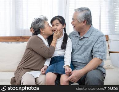 Happy asian grandparents sitting on sofa at home playing and kiss on cheek of little granddaughter