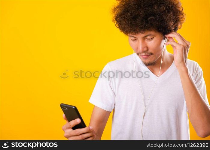 Happy asian handsome man with curly hair he smiling enjoying listening to music on earphones using a mobile smartphone isolate on yellow background