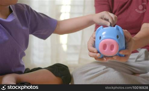 Happy Asian kid boy preschool and mother putting pin money coins into piglet. Little child putting coin into piggy bank for saving with pile of coins at home, Investment education concept,