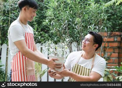 Happy Asian LGBTQ gay men couple wear apron shoveling watering a plant pot outdoors at home. Young man smiling cheerfully, giving a pot plant to boyfriend. Lgbtq lifestyle, love, valentine day concept