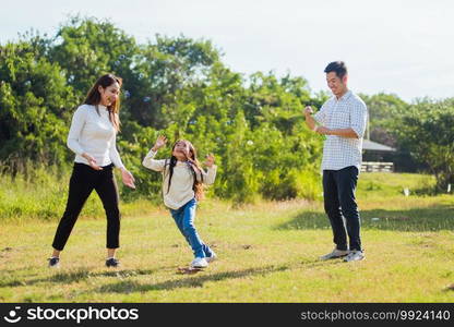 Happy Asian lifestyle family mother, father and little cute girl child having fun together and enjoying outdoor play blowing soap bubbles in the garden park on a sunny day, summertime