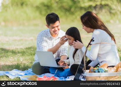 Happy Asian lifestyle young family father, mother and little girl having fun outdoor sitting on picnic blanket using laptop computer technologies while relaxing to open song at garden park in summer