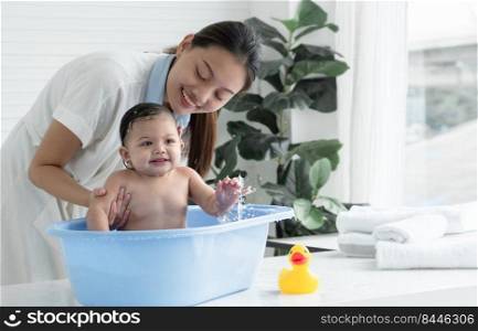 Happy Asian little baby sitting and enjoy playing water in bathtub while young mother wear bathrobe is bathing her cute daughter at home. Baby bathing concept.