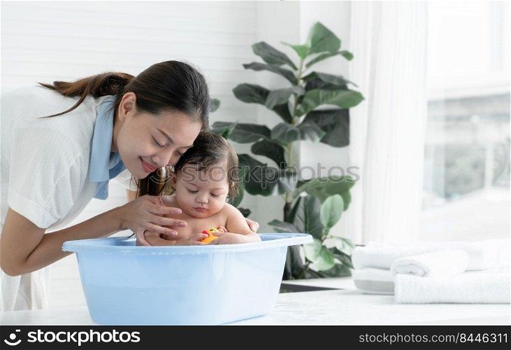 Happy Asian little baby sitting and enjoy playing yellow duck toy in bathtub while young mother wear bathrobe bathing her cute daughter at home. Baby bathing concept. White background
