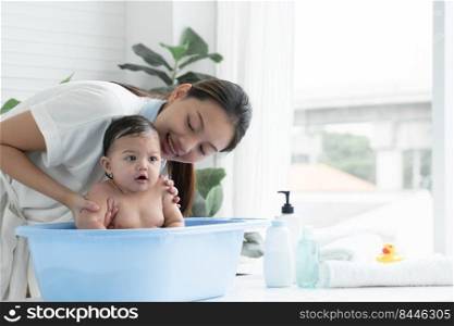 Happy Asian little baby smiling, sitting and enjoy playing water in bathtub while young mother wear bathrobe is bathing her cute daughter at home. Baby bathing concept
