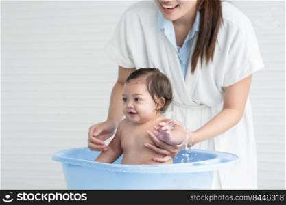 Happy Asian little baby smiling, sitting and enjoy playing water in bathtub while young mother wear bathrobe bathing her cute daughter at home. Baby bathing concept. White background