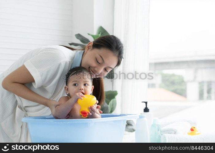 Happy Asian little baby smiling, sitting and enjoy playing yellow duck toy in bathtub while young mother wear bathrobe is bathing her cute daughter at home. Baby bathing concept