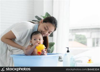 Happy Asian little baby smiling, sitting and enjoy playing yellow duck toy in bathtub while young mother wear bathrobe is bathing her cute daughter at home. Baby bathing concept