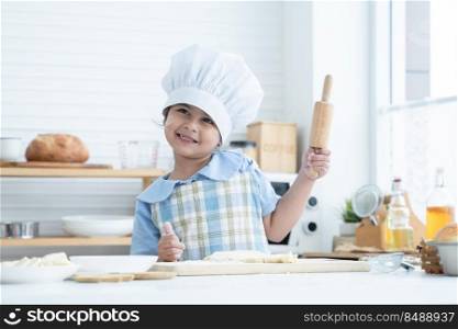 Happy Asian little cute kid girl wear chef hat and apron holding wooden rolling pin and thumbs up while knead dough bread in kitchen at home or cooking class at school. Child education concept