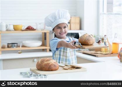 Happy Asian little cute kid girl wear chef hat and apron holding showing homemade whole grains bread on wooden tray in kitchen at home or cooking class at school. Child education concept