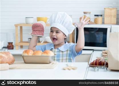 Happy Asian little cute kid girl wear chef hat, apron and oven glove proud with homemade bread on loaf pan finished from oven in kitchen at home or cooking class at school. Child education concept