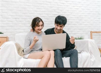 Happy Asian Lover sitting and using the digital laptop in cheerful action over the sofa of living room at modern home, Couple successfull and lifestyle concept
