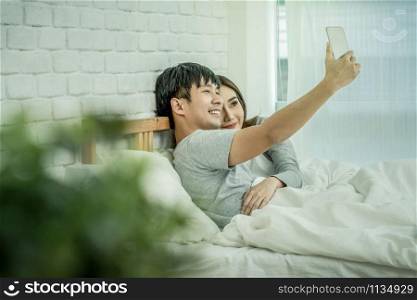 Happy Asian Lover sitting and using the digital smart mobile phone to selfie on the bed of bedroom at modern home, Couple successfull and lifestyle concept