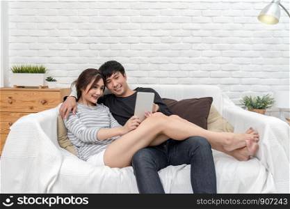 Happy Asian Lover sitting and using the digital technology tablet to selfie over the sofa of living room at modern home, Couple successfull and lifestyle concept