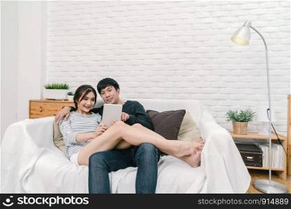 Happy Asian Lover sitting and using the digital technology tablet to selfie over the sofa of living room at modern home, Couple successfull and lifestyle concept