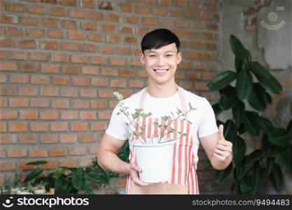 Happy Asian man wear apron holding a plant pot outdoors at home. Young healthy man smiling cheerfully, standing, thumbs up while taking care of plant at garden backyard