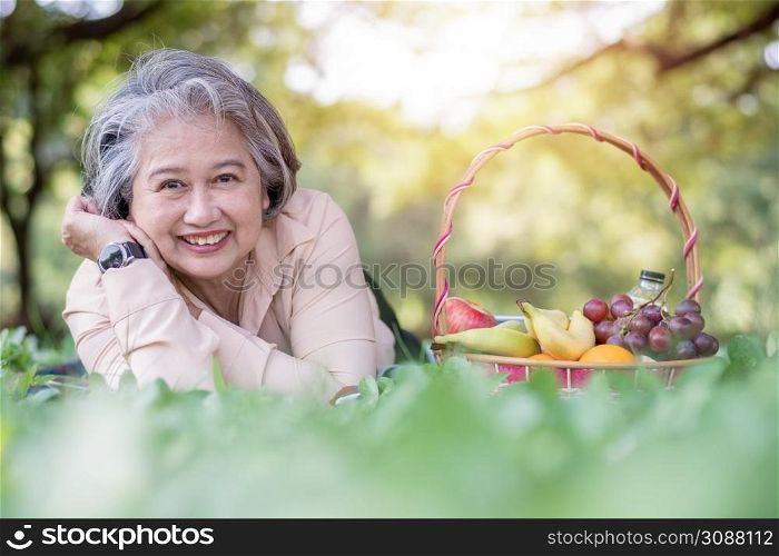 Happy Asian old senior woman and wear a health watch and lying on the picnic mat in park and basket of fruit besides. Concept of happy elderly woman after retirement and good health