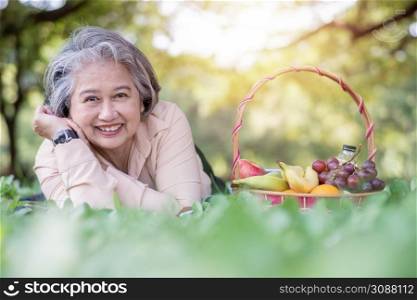 Happy Asian old senior woman and wear a health watch and lying on the picnic mat in park and basket of fruit besides. Concept of happy elderly woman after retirement and good health