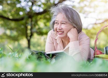 Happy Asian old senior woman and wear a health watch and reading book and lying on the picnic mat in park and basket of fruit besides. Concept of happy elderly woman after retirement and good health
