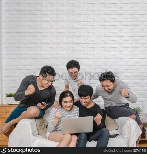 Happy Asian people group sitting and using the digital laptop in cheerful and celebration action over the sofa of living room at modern home, Couple successfull and lifestyle concept