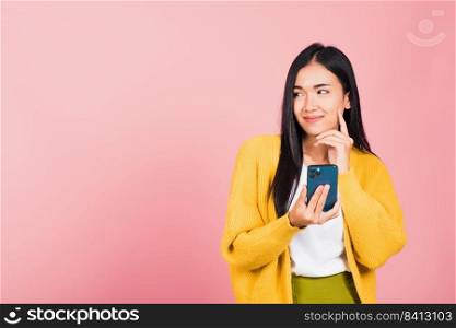 Happy Asian portrait beautiful cute young woman excited smiling holding mobile phone and think idea finger touch face, studio shot isolated on pink background, female using smartphone making gesture