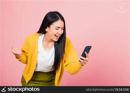 Happy Asian portrait beautiful cute young woman teen smiling excited using mobile phone say yes! Studio shot isolated on pink background, Thai female surprised make winner gesture on smartphone