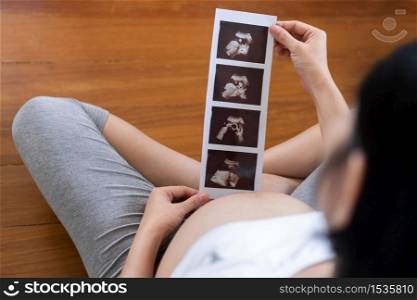 Happy Asian Pregnant woman looking ultrasound image while touch her belly at home. Young Mother holding sonogram of her unborn baby. Concept of pregnancy, Maternity prenatal care. Mom with a new life