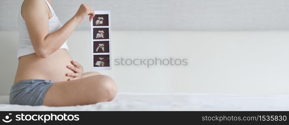 Happy Asian Pregnant woman sitting holding ultrasound image while touch her belly on bed. Mother with sonogram of her unborn baby in white room, Banner, Concept of pregnancy, Maternity prenatal care