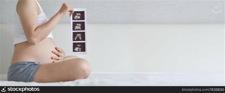Happy Asian Pregnant woman sitting holding ultrasound image while touch her belly on bed. Mother with sonogram of her unborn baby in white room, Banner, Concept of pregnancy, Maternity prenatal care