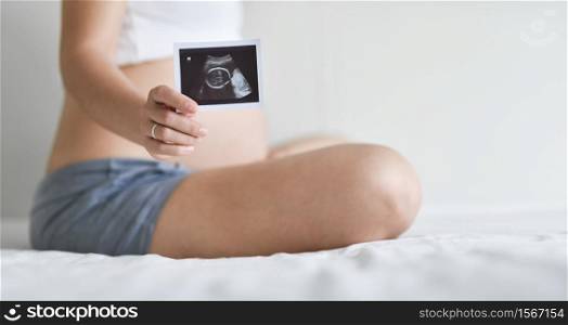 Happy Asian Pregnant woman sitting showing ultrasound picture while sitting on bed. Mother with sonogram of her unborn baby in white room, Concept of pregnancy, Maternity prenatal care
