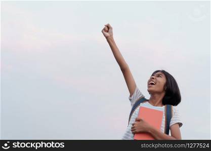 Happy Asian schoolgirl success and excited on the face while holding a book with hand raised up to blue sky background in the summer at morning