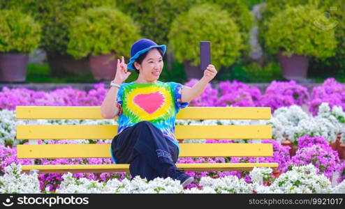 Happy Asian smiling female tourist showing love hand sign while taking selfie or video call with smartphone on park bench in pink and white bougainvillea field of Nong Nooch tropical garden, Thailand