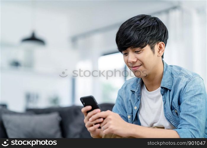 Happy asian teenager using smart phone and smiling on sofa living room at home. Asian man holding and using cellphone for searching data and social medie on internet. Teenager and Technology concept.
